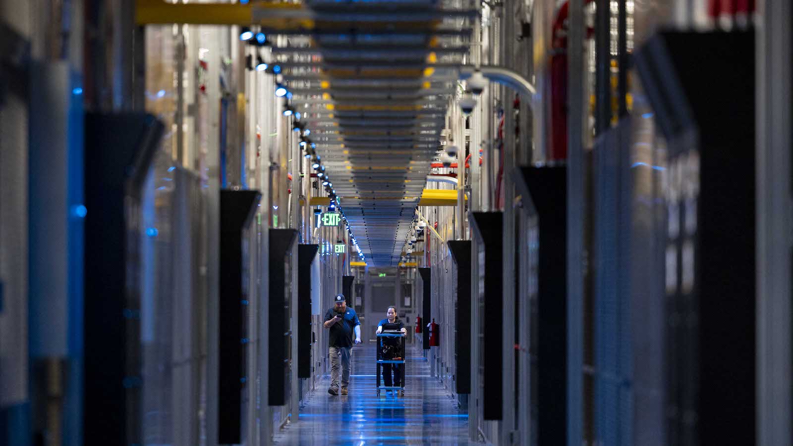 Two people walking down a hallway lit with blue lights and large looming computer towers.