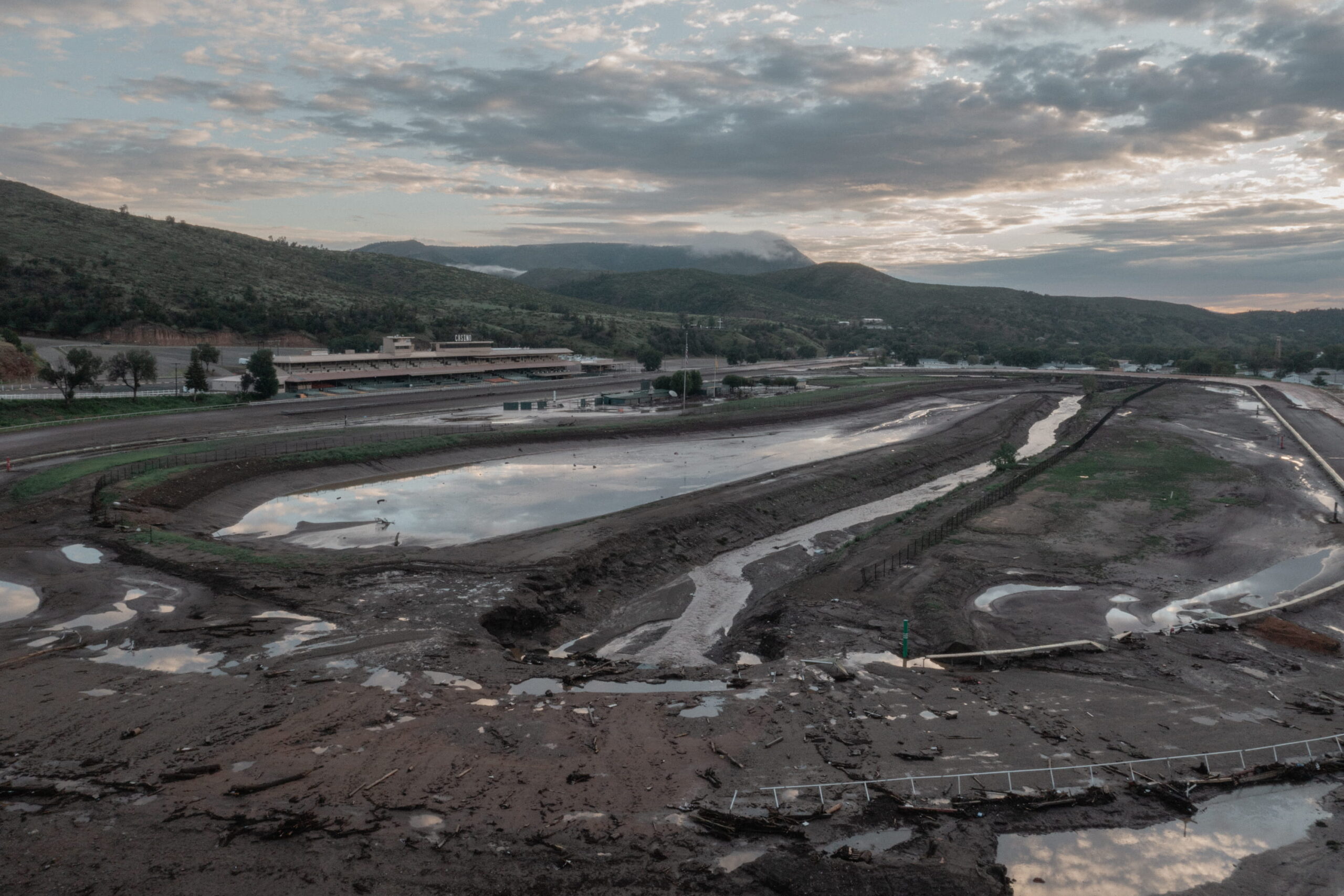An elevated view shows a mud-caked, flooded horse-racing track surrounded by standing water and mountain ridges under a cloudy sky.