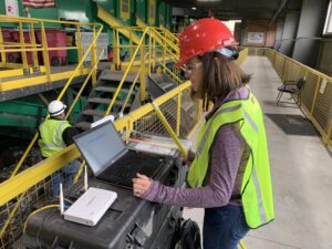 Susan Graff with brown hair wearing a res hard hat and reflective vest looking at a computer in a material recovery facility