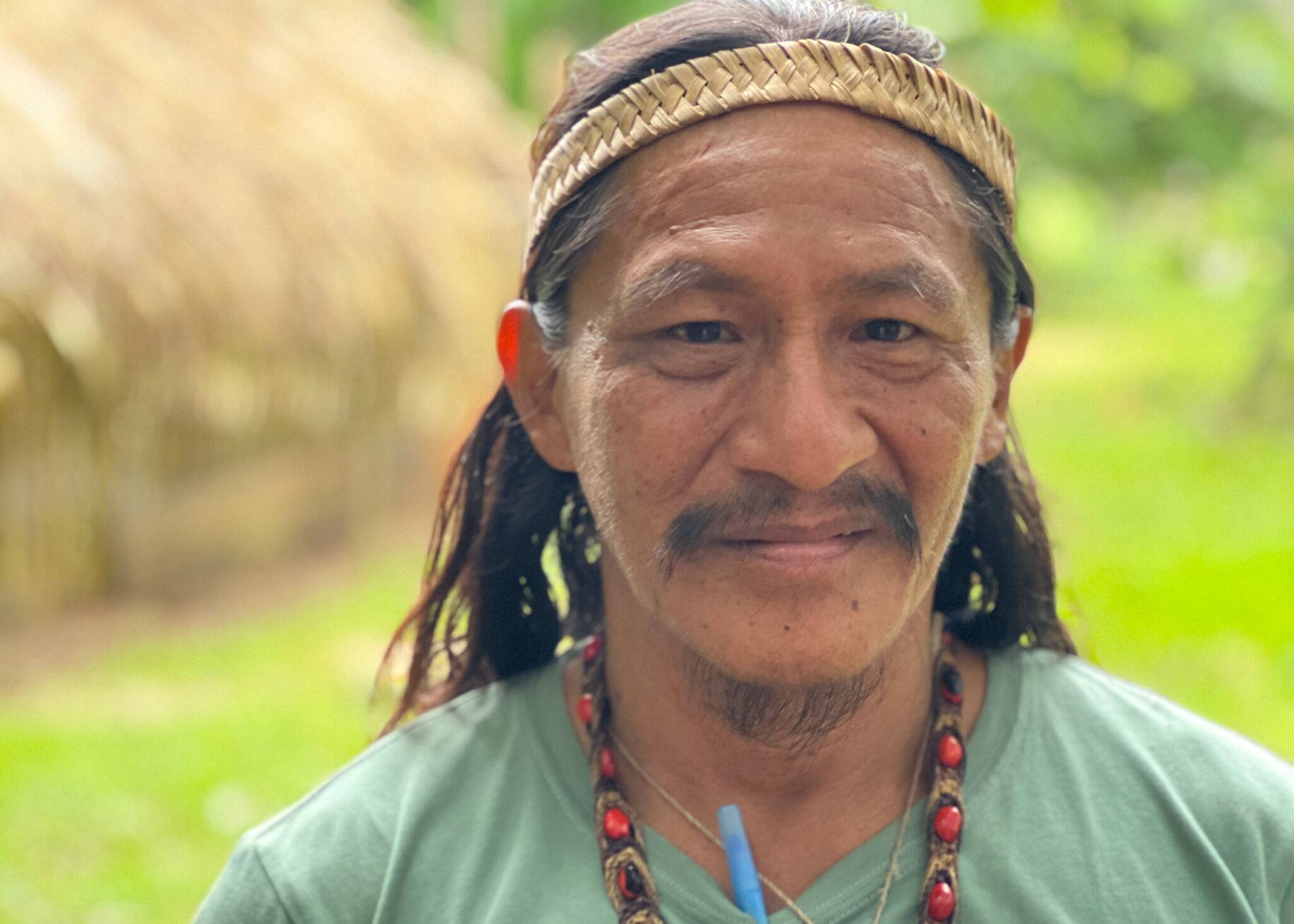 A mustachioed man in a green shirt and red necklace with a reed headband smiles