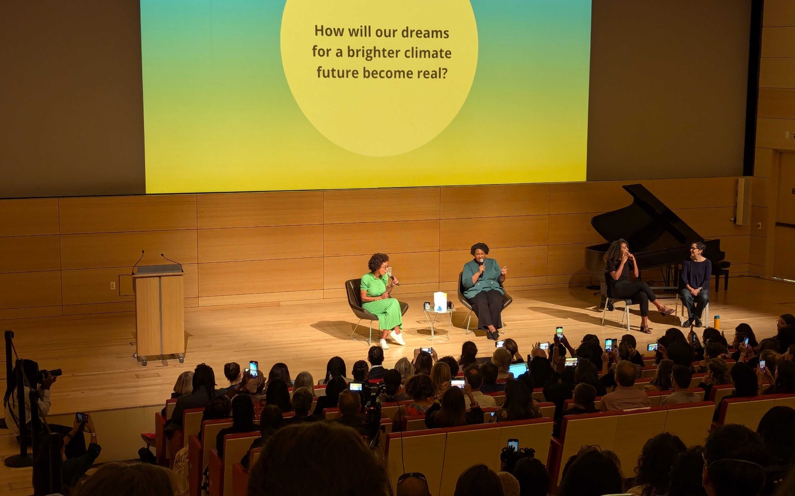 Two women sit on a warmly lit stage holding microphones, in front of an audience of people holding up their phones to record