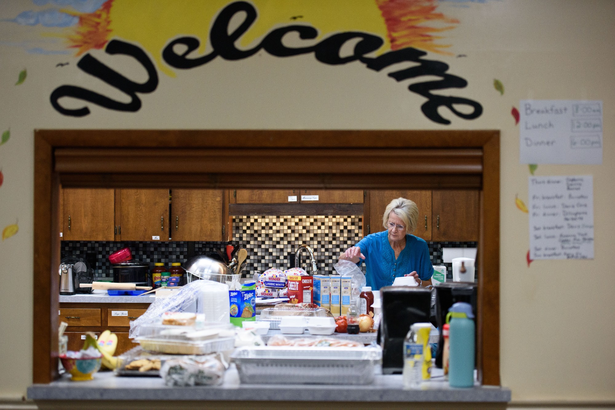 Visible through a small window with the word Welcome above it, a woman stands in a kitchen behind an array of food including a loaf of bread, boxes of crackers, and Ziploc baggies.