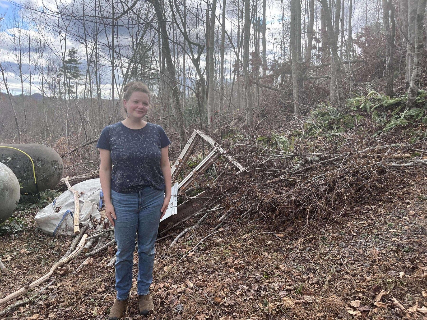 A blonde woman in jeans and a blue t-shirt stands in a bare forest in front of a pile of sticks and debris.