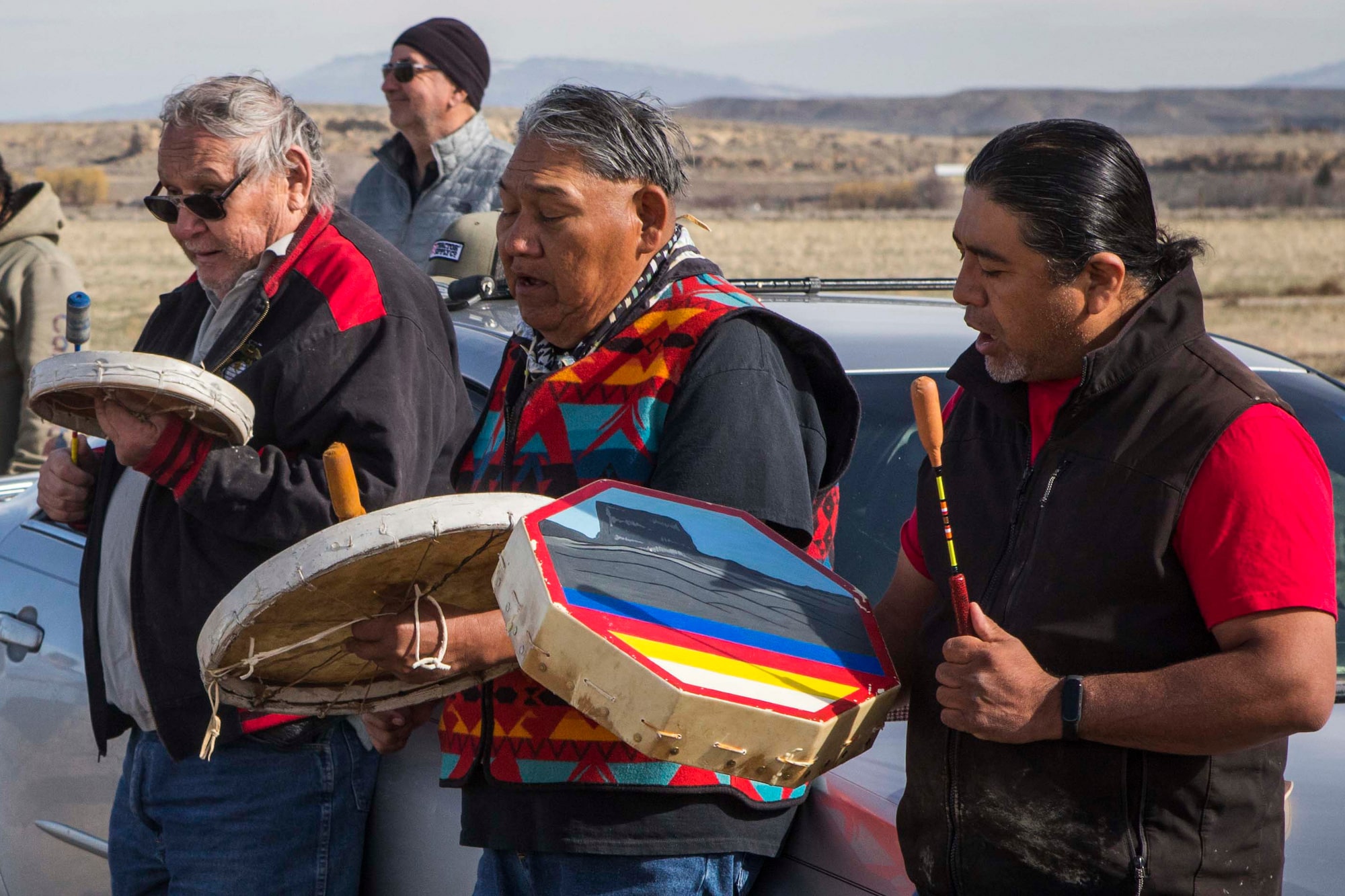 A group of people hold drums and sing while standing on a plain