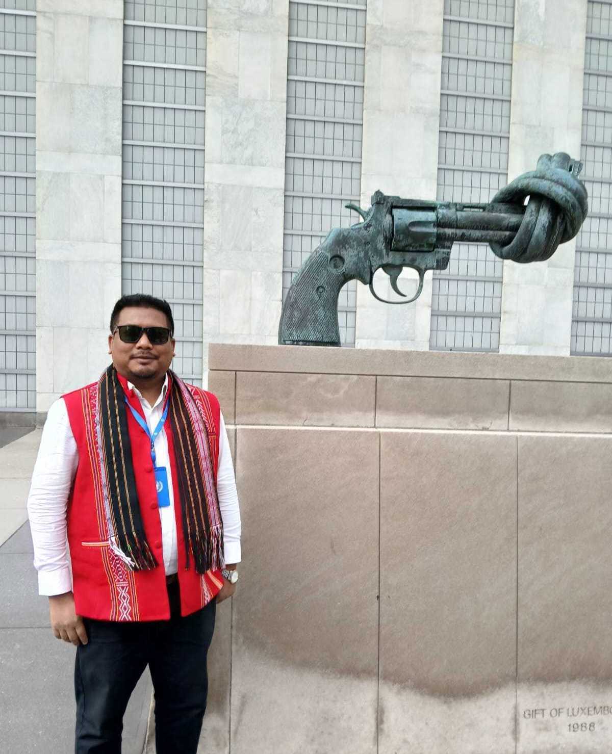 A man in a red vest stands next to a statue of a gun with a muzzle tied up so it can't fire