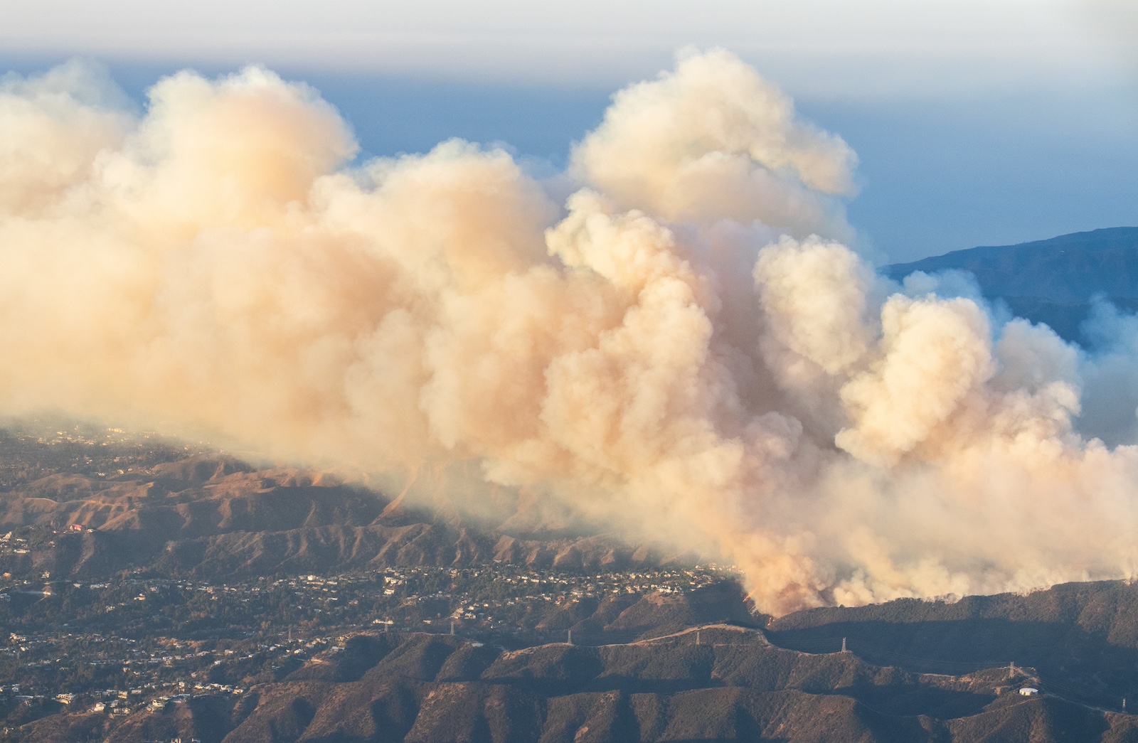 An aerial view of a huge plume of tan smoke emerging from partially developed mountainous terrain