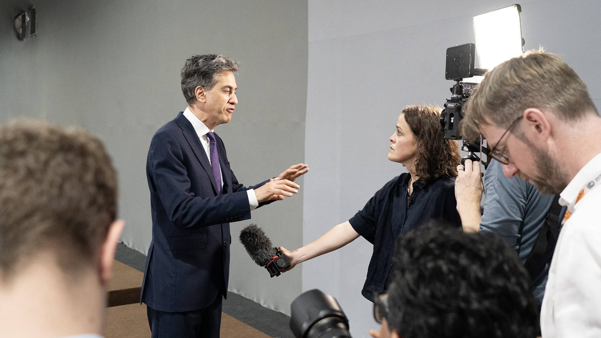Edward Miliband, wearing a black suit and a purple tie, speaks to a team of reporters holding a microphone and a light at COP30 in Brazil.
