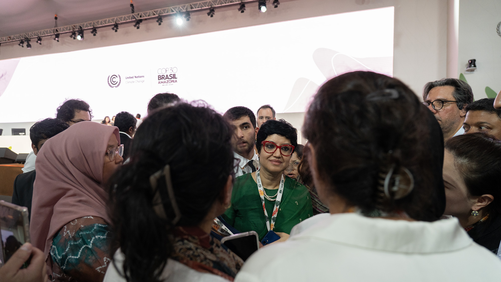 Suman Chandra, wearing a green shirt and red glasses, speaks in the middle of a crowd of negotiators at COP30 in Brazil.