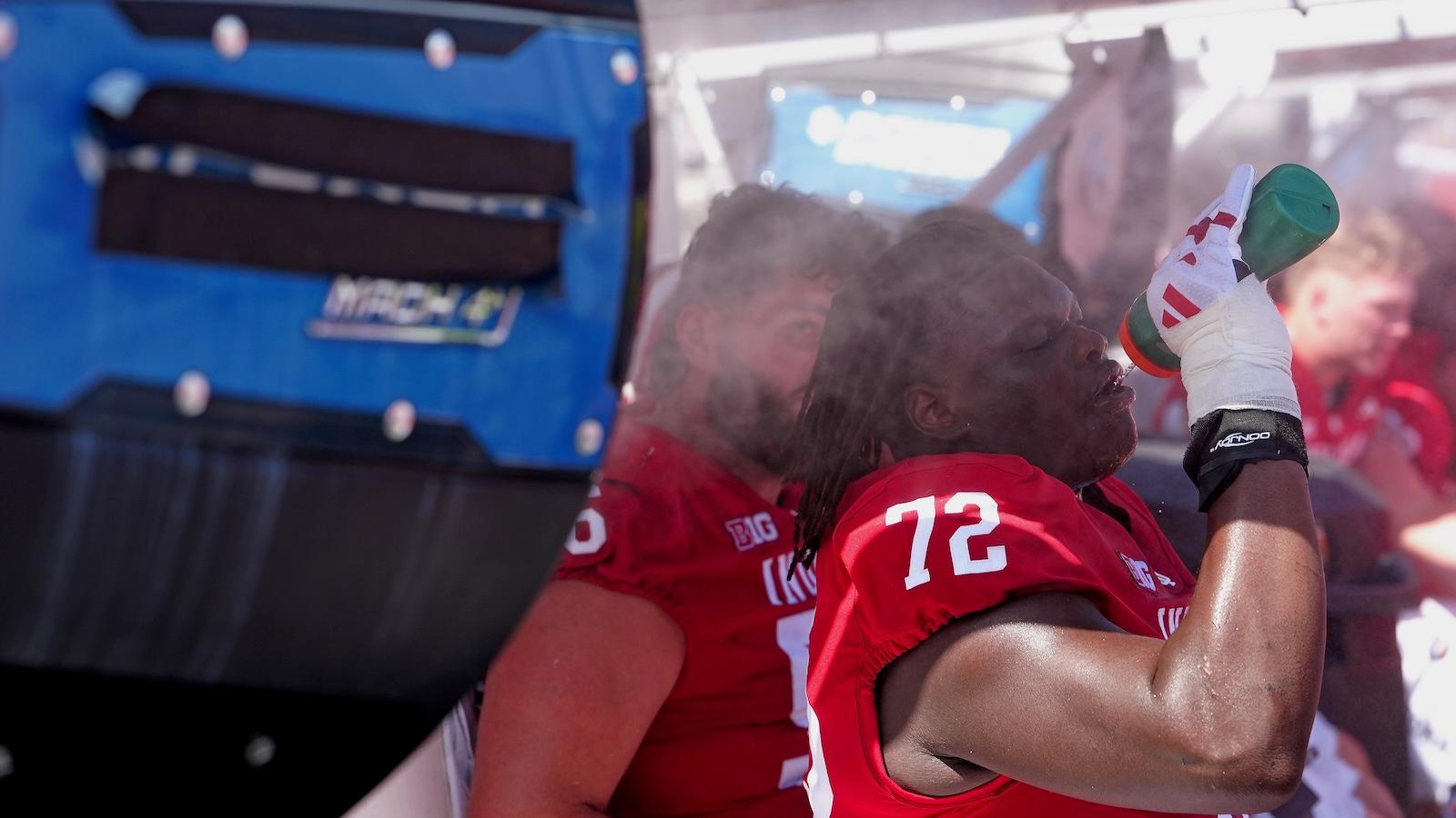 Two college football players sit in front of a large fan and sip water as they attempt to keep cool during a game.
